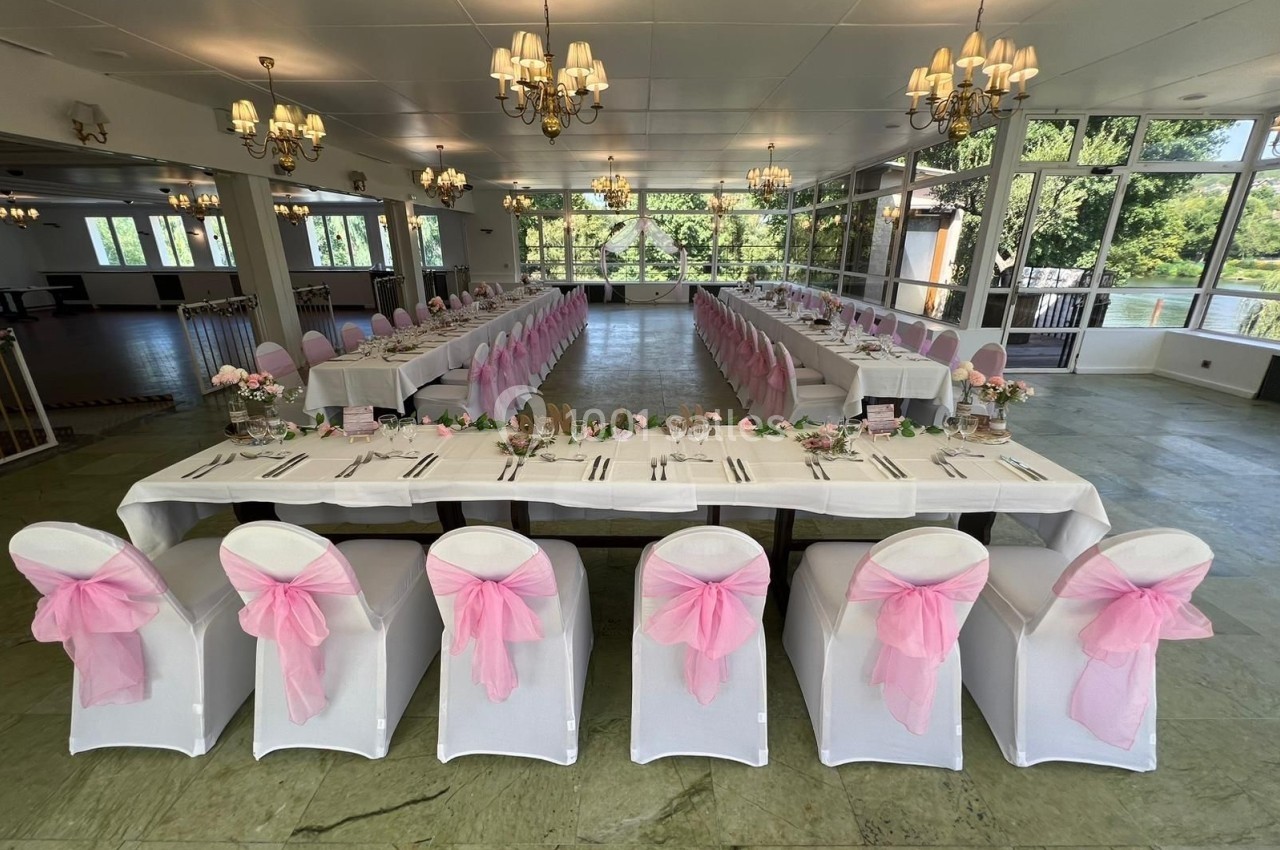 Salle de réception lumineuse avec deux longues tables décorées de nappes blanches et de chaises ornées de nœuds roses.