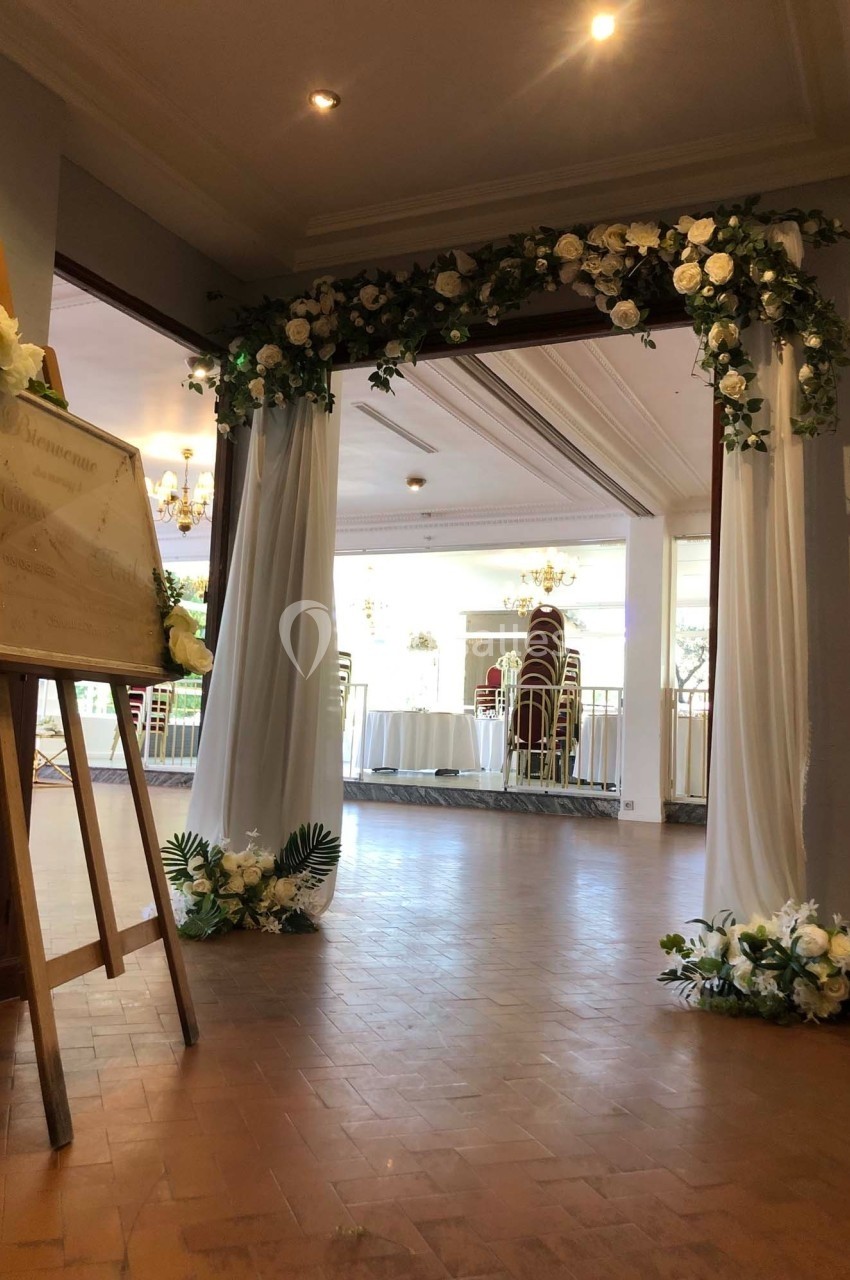 Arche décorée de fleurs blanches et feuillages encadrant l'entrée d'une salle lumineuse avec des tables et chaises.