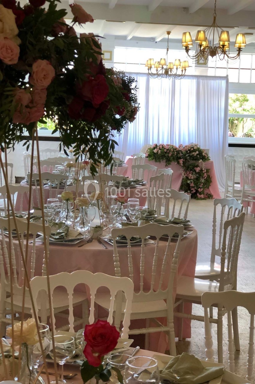 Salle de réception décorée avec des tables rondes, nappes roses, chaises blanches et arrangements floraux élégants.