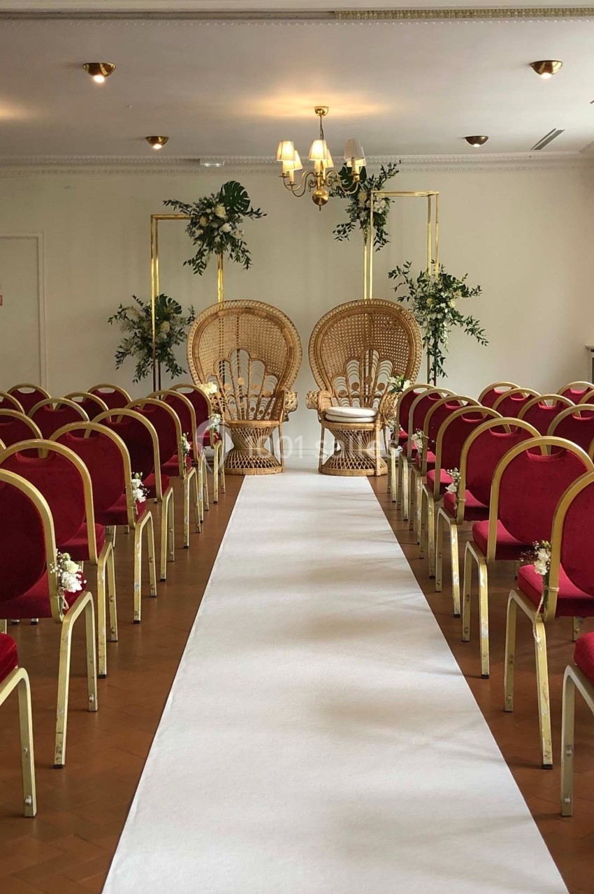 Salle de cérémonie avec allée centrale blanche, chaises rouges alignées et deux fauteuils en rotin décorés de plantes.