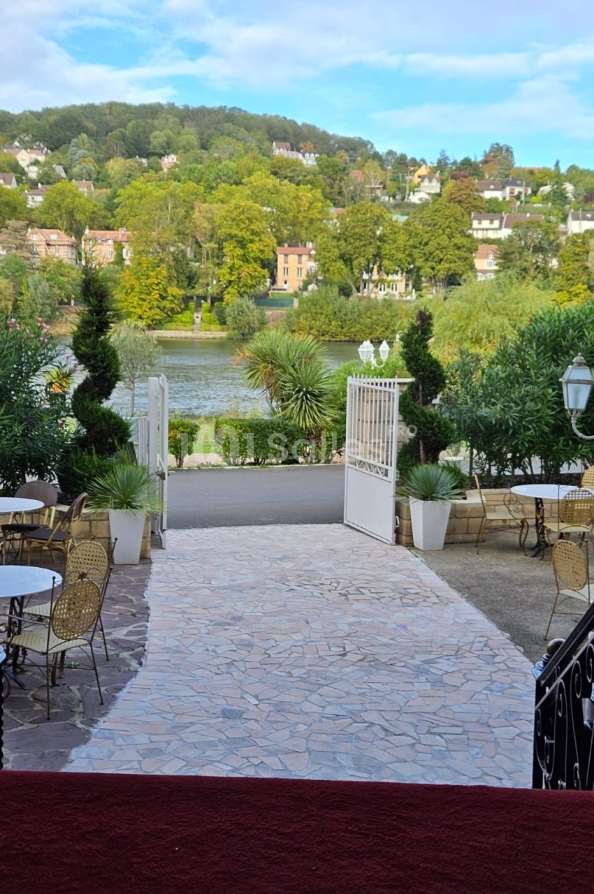 Vue sur une terrasse avec des tables et des chaises, donnant sur un jardin, une rivière et des collines verdoyantes.