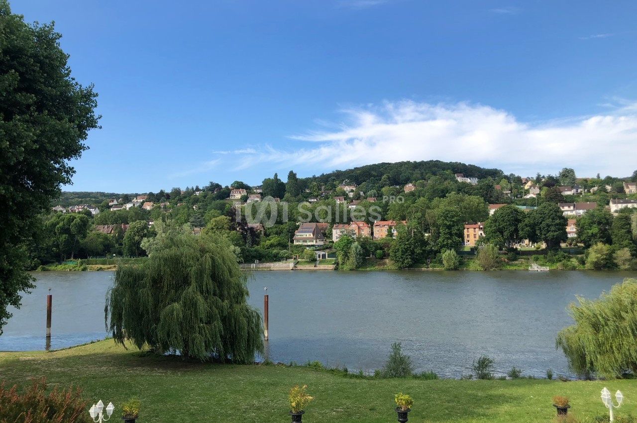 Vue d'une rivière bordée de verdure avec des collines boisées et des habitations en arrière-plan sous un ciel dégagé.