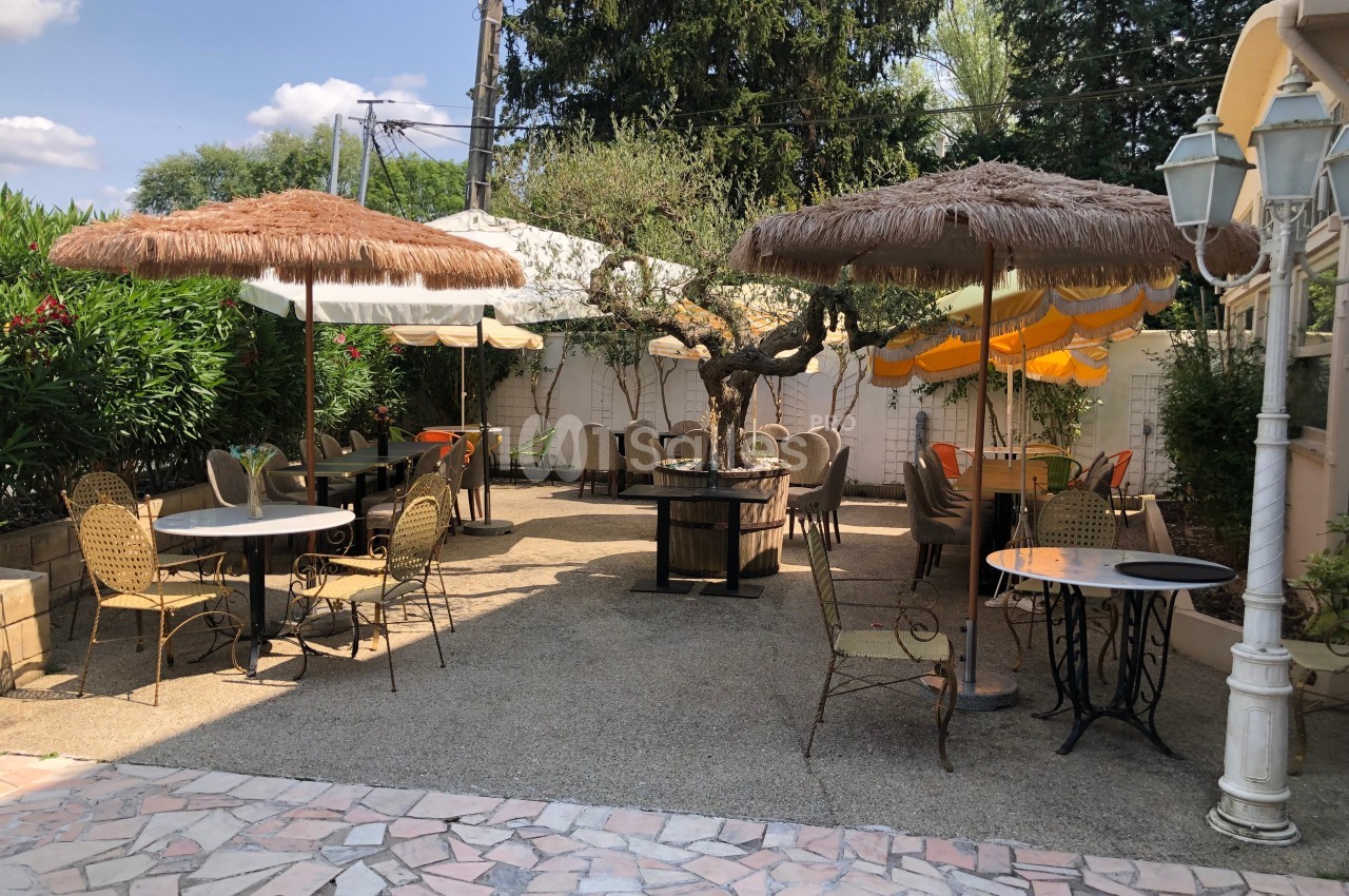 Terrasse extérieure avec tables, chaises en osier et parasols en paille, entourée de végétation et d'arbres.