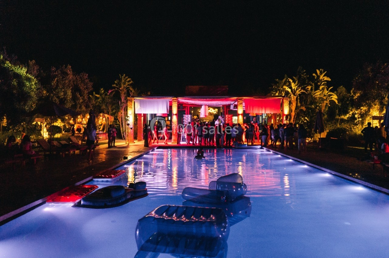 Soirée festive autour d'une piscine éclairée, avec des invités rassemblés devant un bâtiment illuminé.
