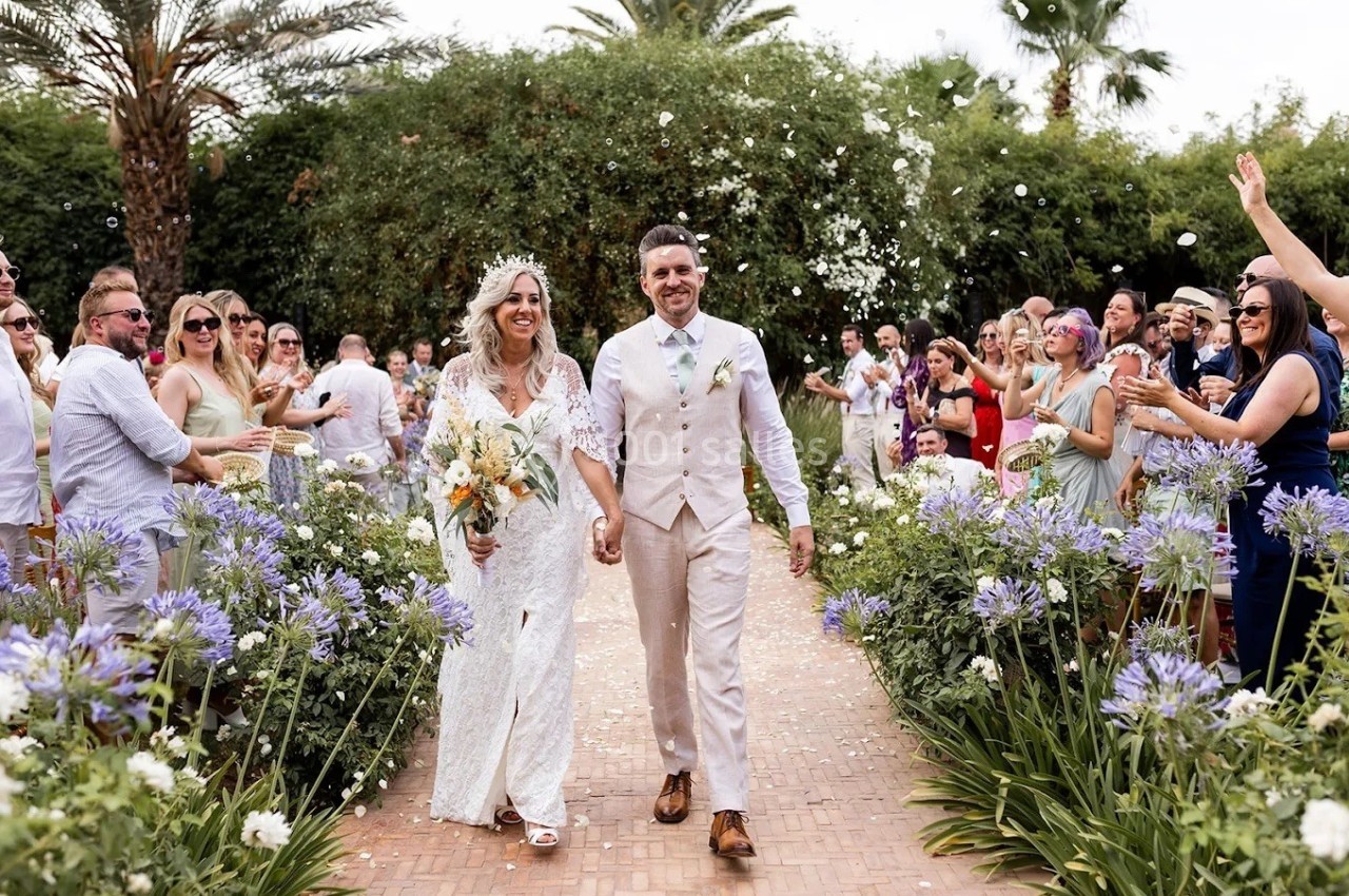 Un couple souriant marche main dans la main lors de leur cérémonie de mariage en extérieur, entouré d'invités.