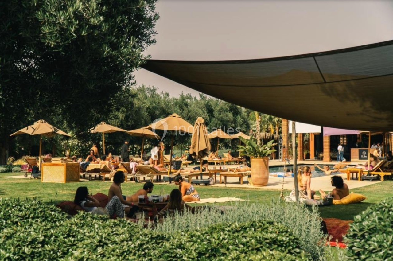 Personnes se relaxant sur des transats et pelouses dans un espace extérieur ombragé avec parasols et végétation.