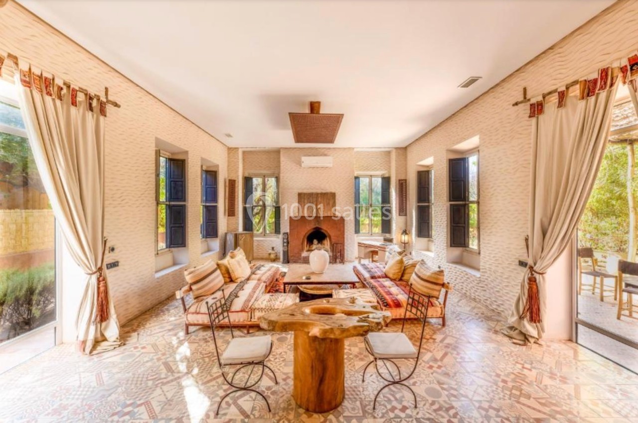 Salon lumineux avec cheminée centrale, mobilier en bois et métal, tapis colorés et grandes fenêtres donnant sur un jardin.