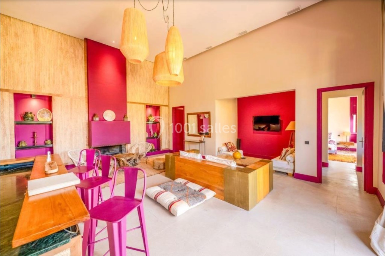 Salon lumineux avec murs rouges, mobilier coloré, cheminée, lampes suspendues et espaces de détente.
