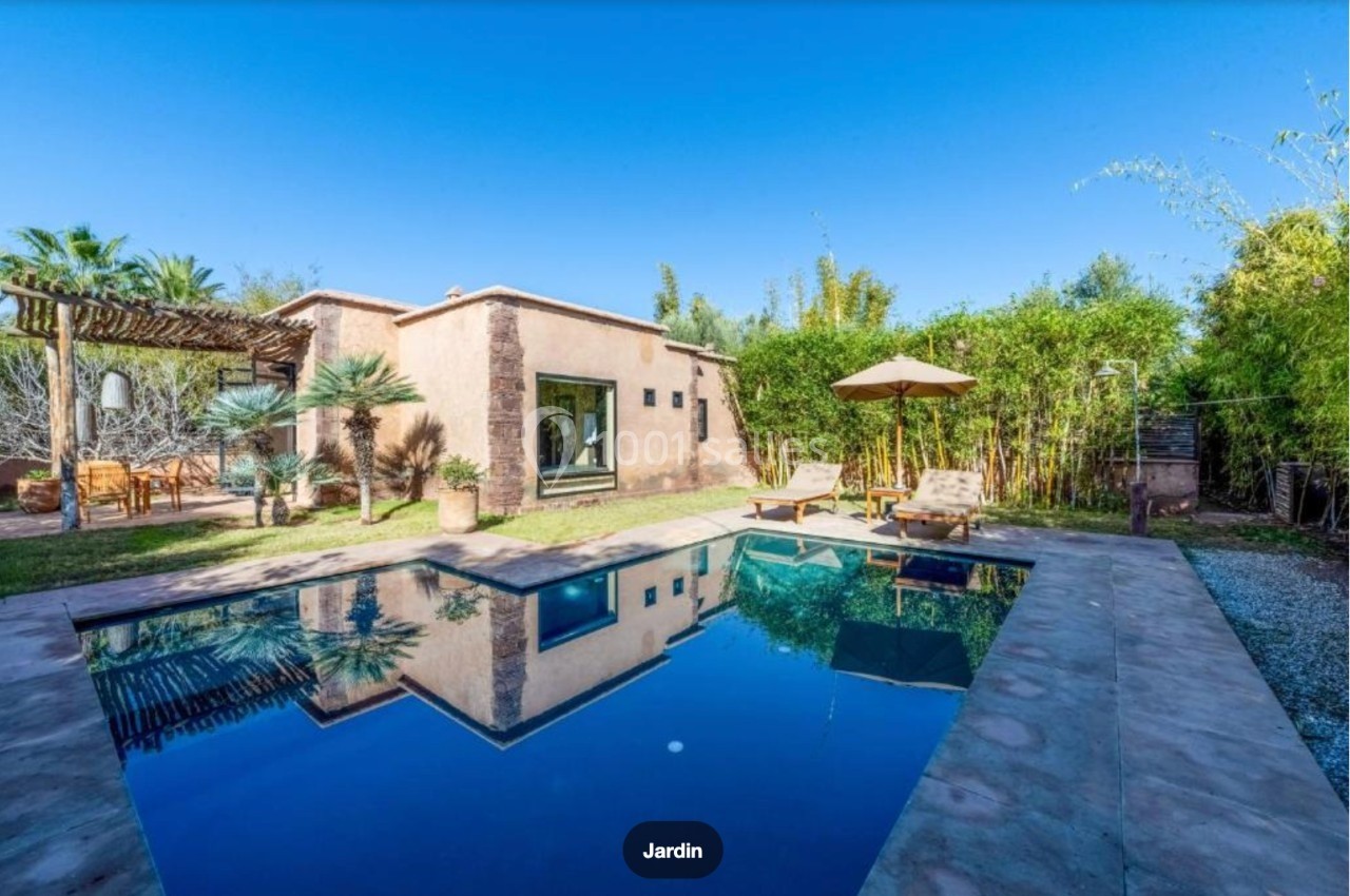 Maison avec piscine entourée de végétation, chaises longues et parasol dans un jardin sous un ciel dégagé.