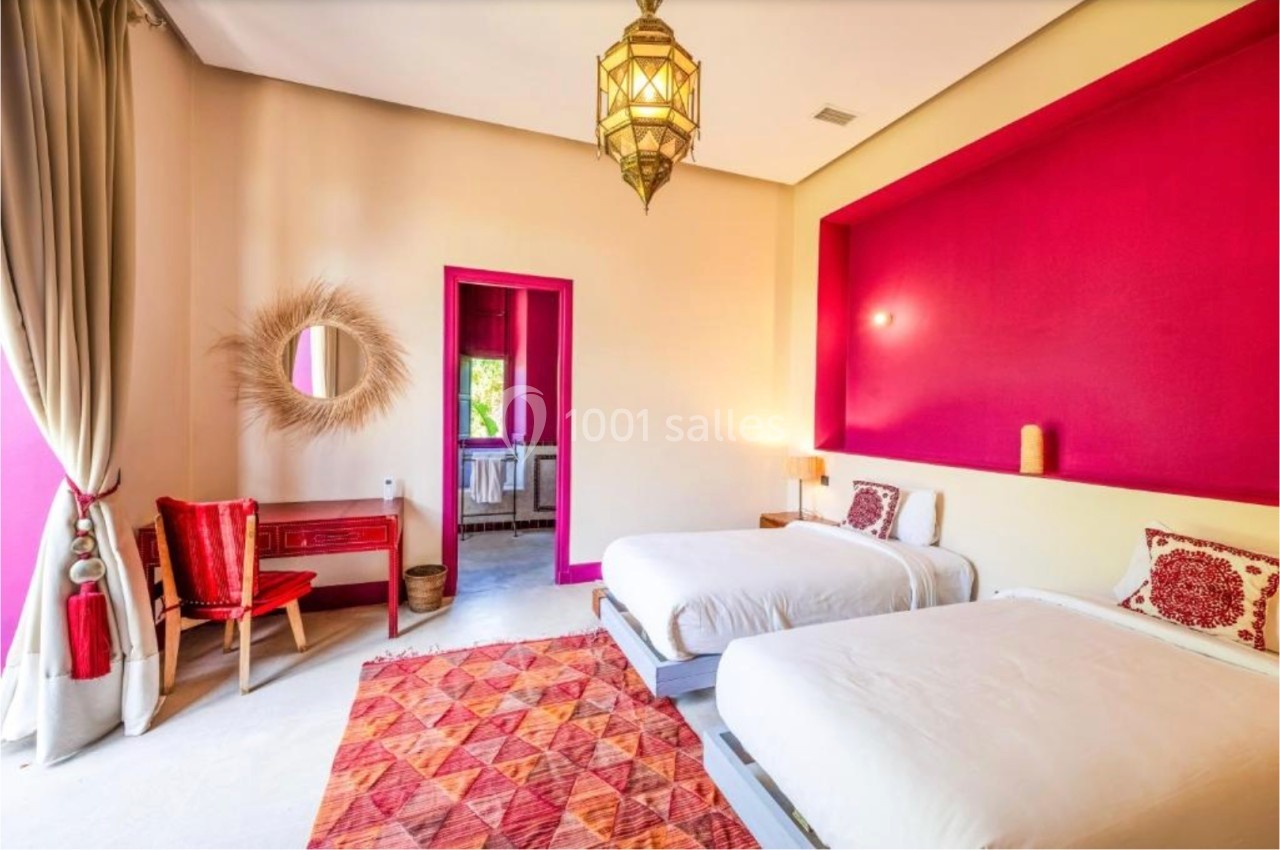 Chambre lumineuse avec deux lits simples, murs rouges et beige, tapis coloré, bureau et miroir décoratif.