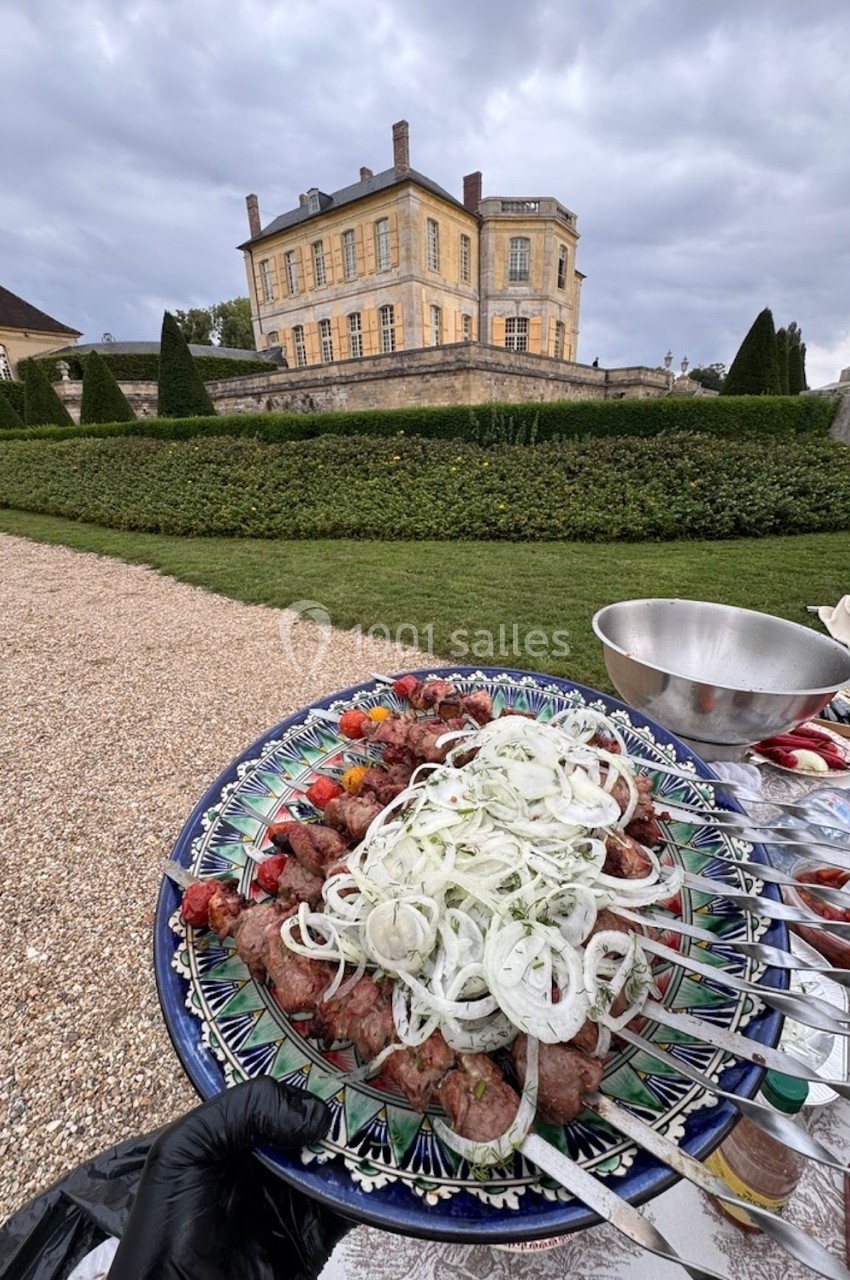 Assiette de brochettes garnies d'oignons et légumes, tenue devant un château entouré de jardins.