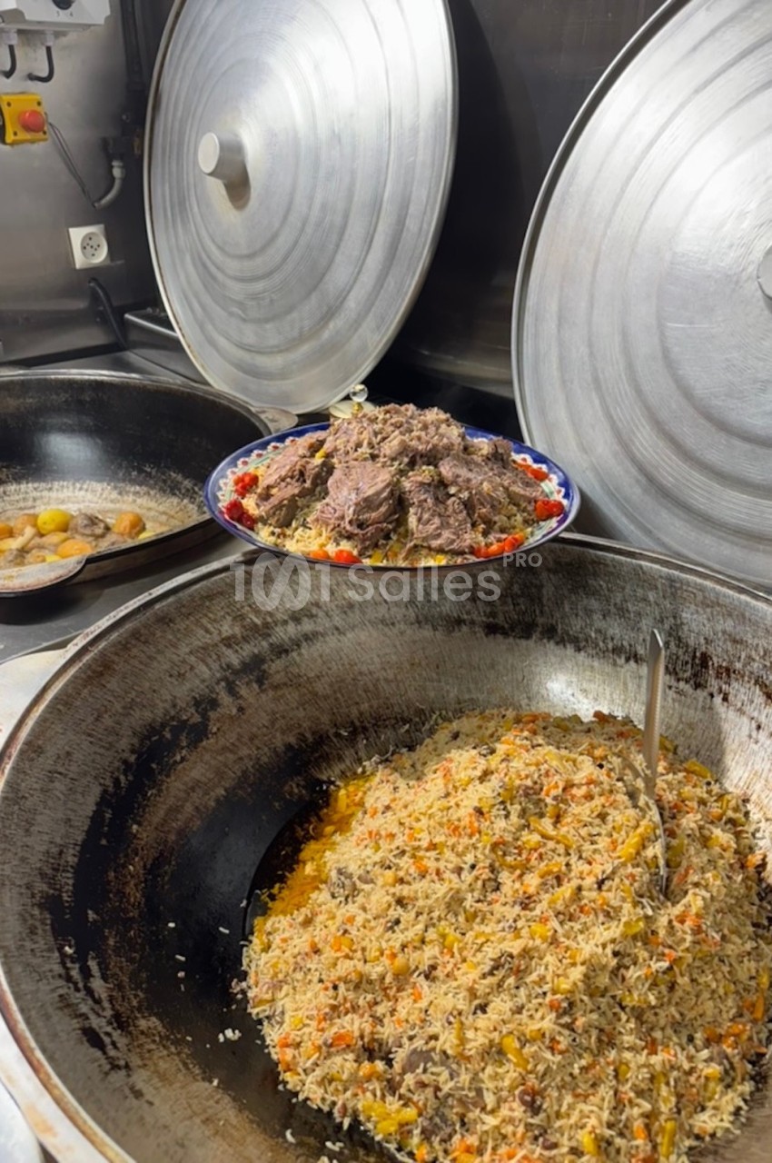 Plat de riz aux légumes et viande dans une grande marmite, avec un plat de viande et légumes en arrière-plan.