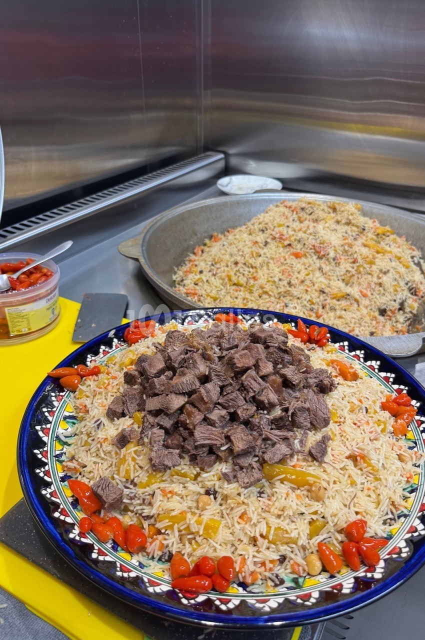 Plat de riz garni de morceaux de viande et de légumes, présenté dans une assiette décorée, avec une poêle en arrière-plan.