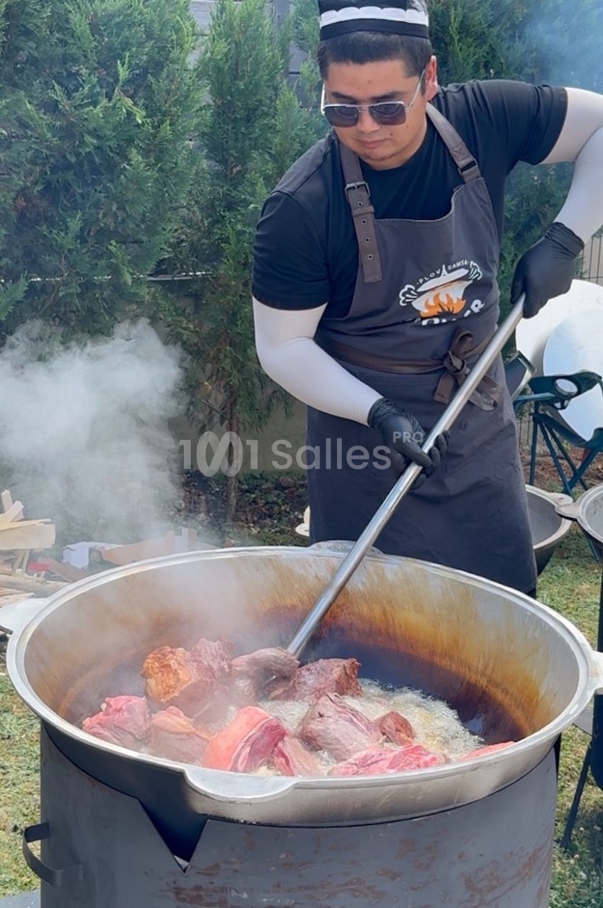 Un homme cuisine de grandes pièces de viande dans une marmite fumante en extérieur, portant un tablier et des gants.
