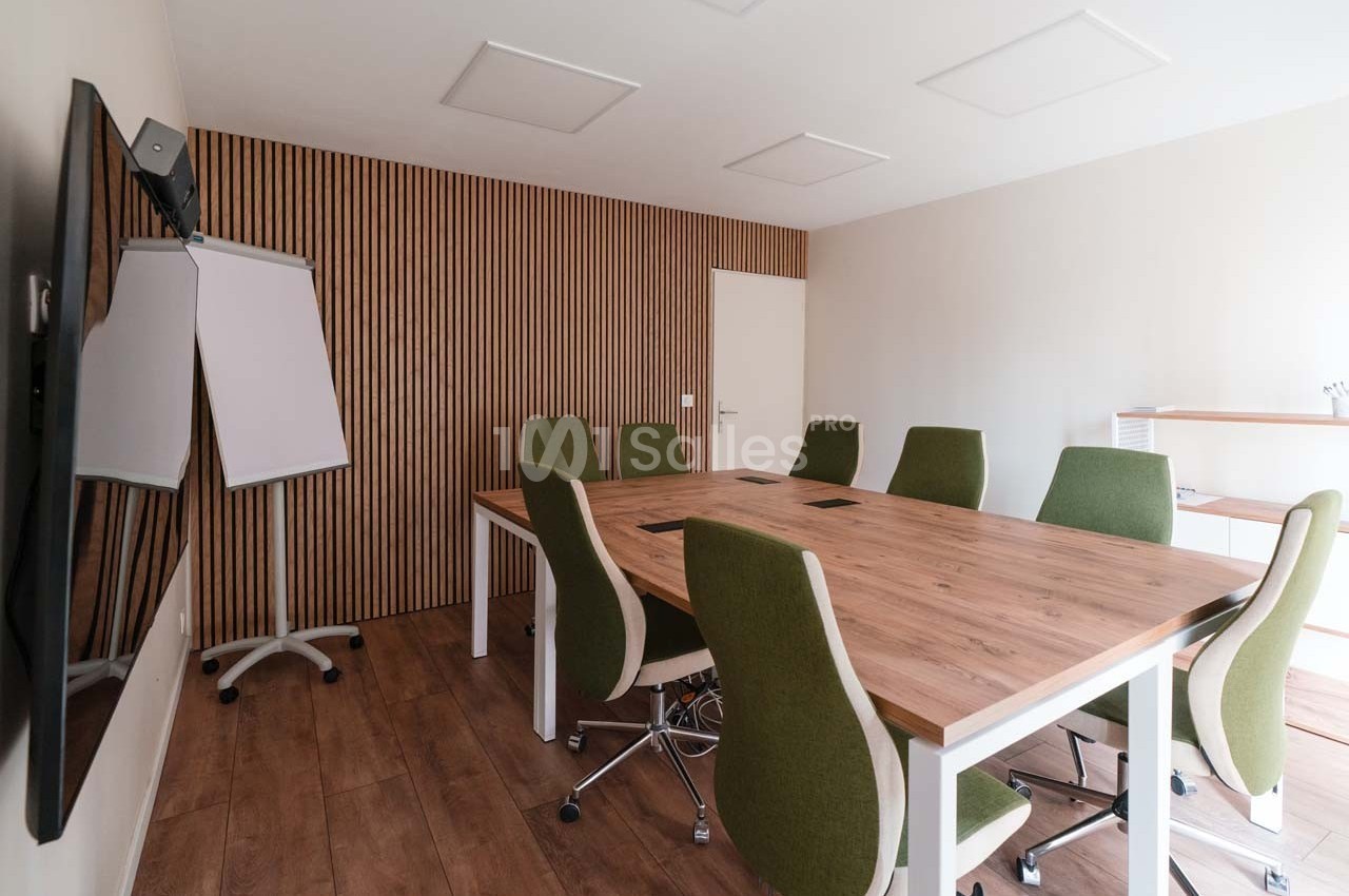 Salle de réunion lumineuse avec table en bois, chaises vertes, tableau blanc et mur en bois à l'arrière.
