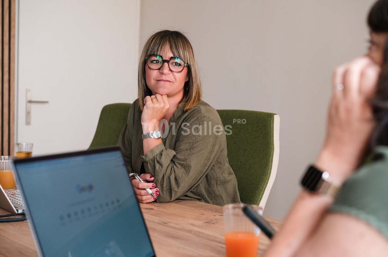 Une femme assise à une table de réunion, regardant devant elle, avec un ordinateur portable et des verres sur la table.