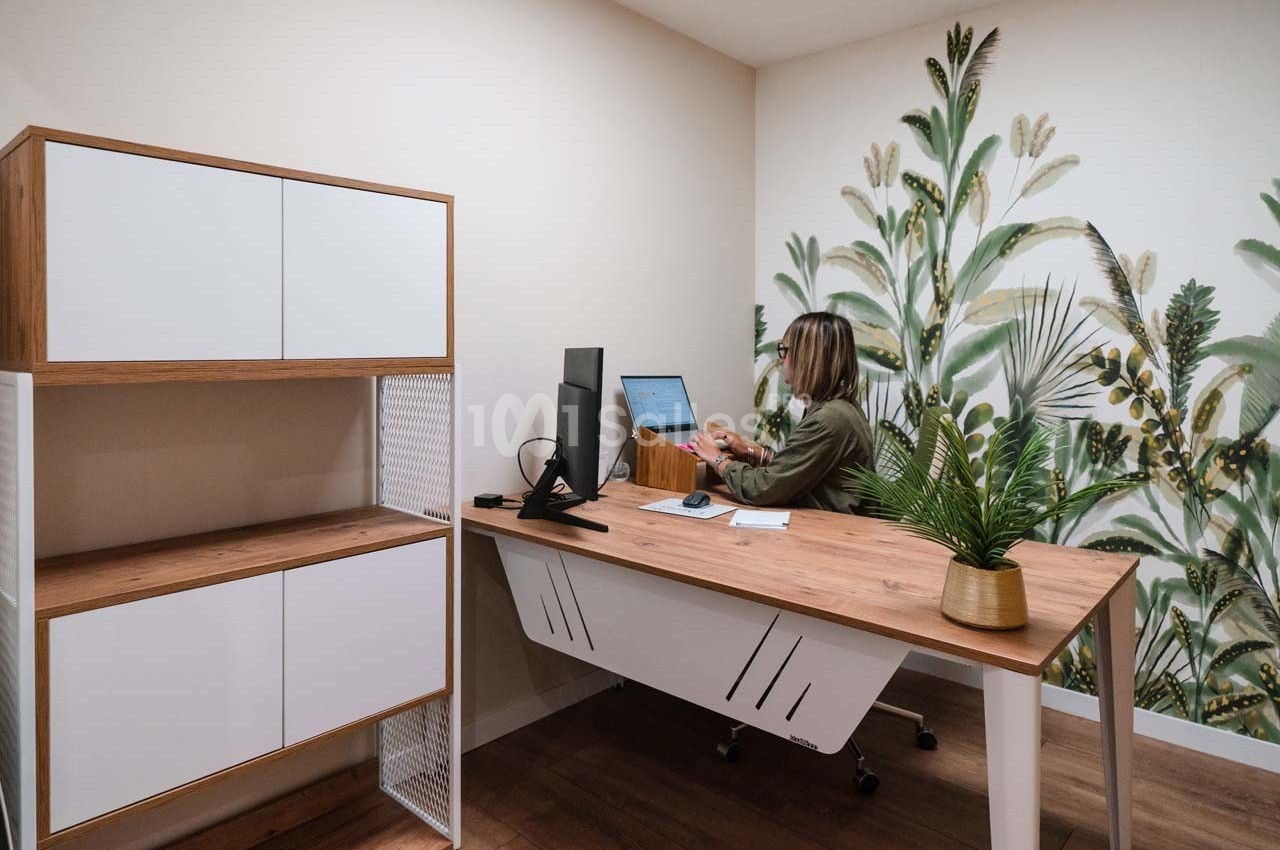 Une femme travaille sur un ordinateur dans un bureau moderne avec un mur décoré de motifs végétaux.