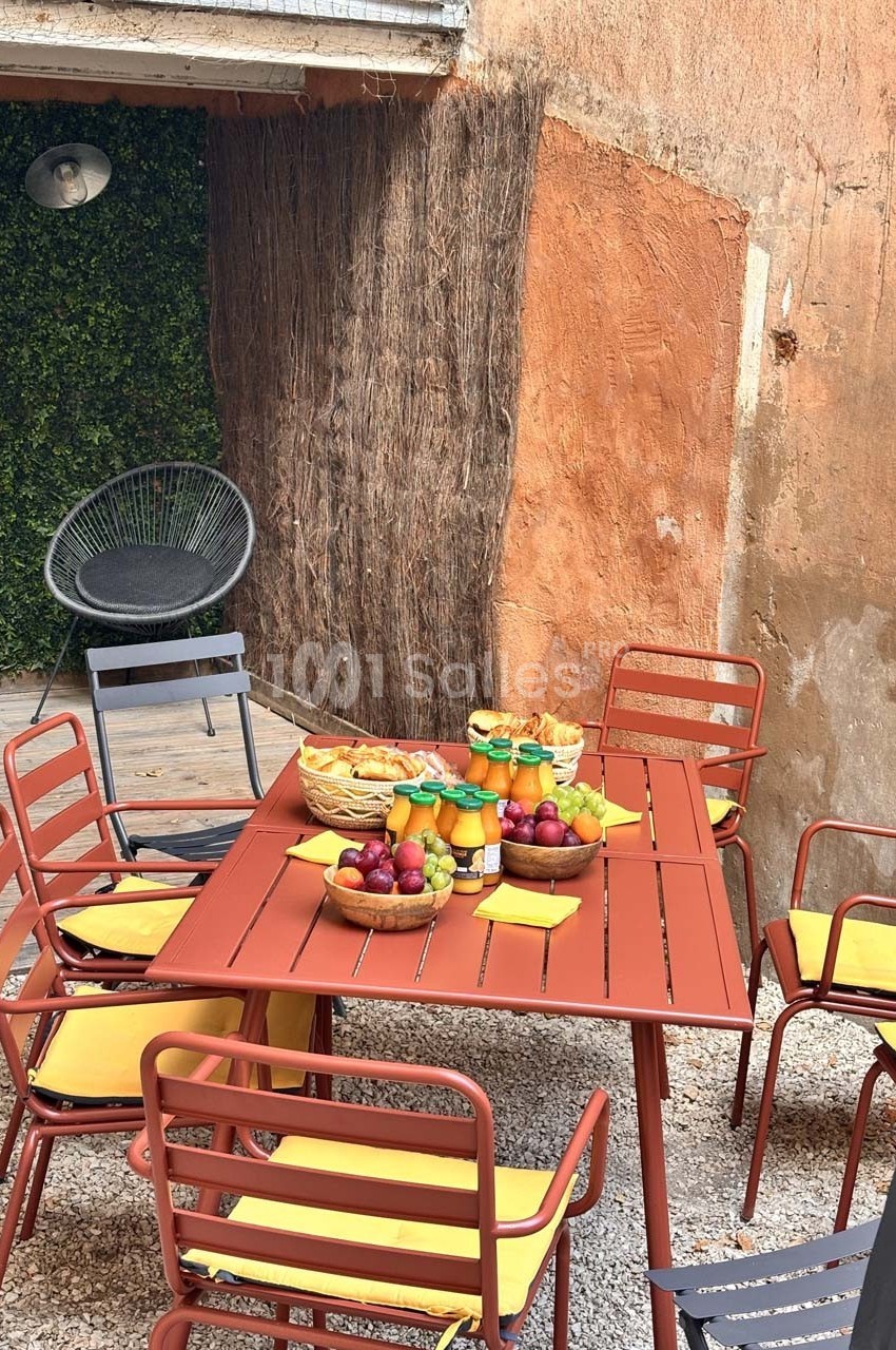 Table extérieure rouge entourée de chaises jaunes, garnie de fruits, jus et pain, dans une cour avec murs en pierre et…