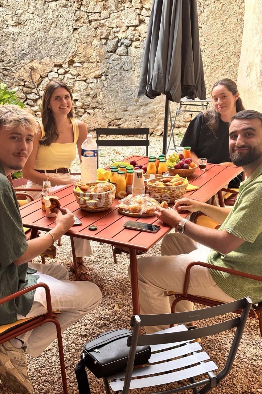 Quatre personnes assises autour d'une table en extérieur, partageant un petit-déjeuner avec fruits, jus et viennoiseries.