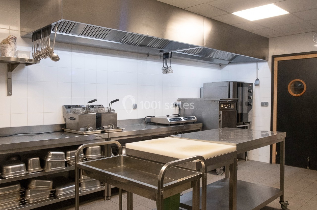Cuisine professionnelle avec plan de travail en inox, équipements de cuisson, étagères et carrelage mural blanc.