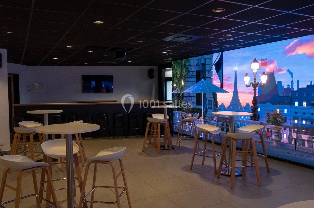 Salle moderne avec tables hautes, tabourets et écran affichant une vue urbaine au coucher du soleil.