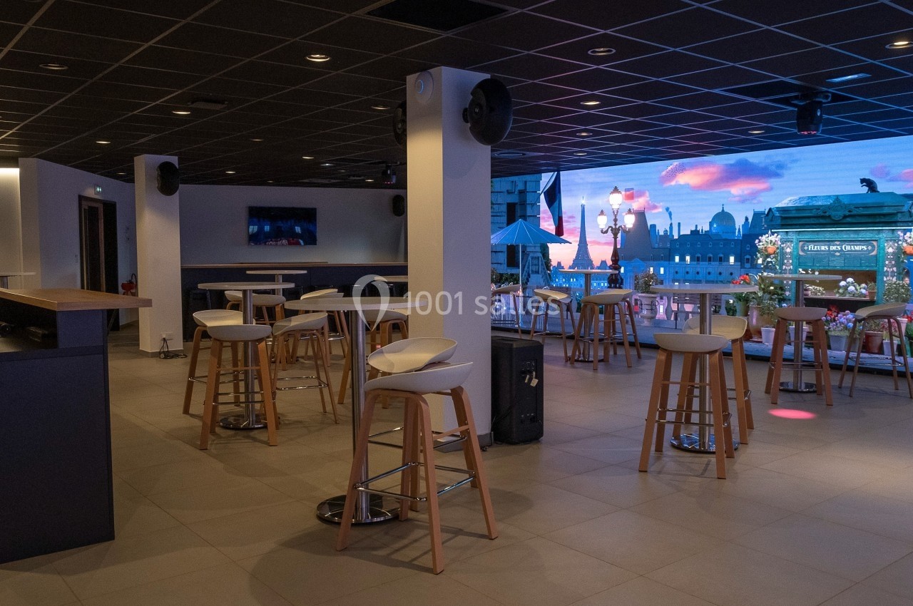 Salle moderne avec tables hautes, tabourets et écran affichant une vue urbaine animée en arrière-plan.