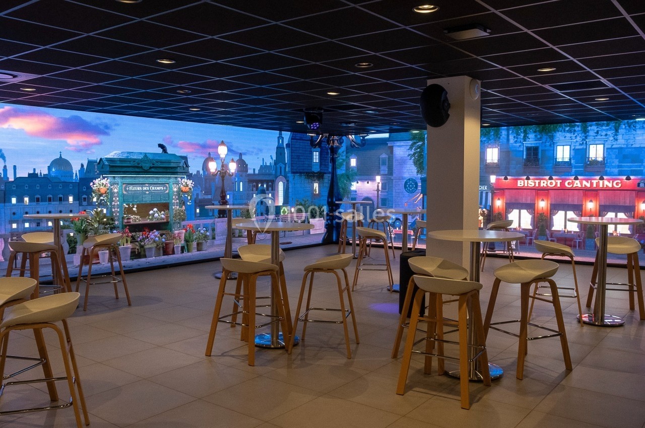 Terrasse intérieure avec tables et tabourets, vue sur une façade lumineuse de restaurant et un décor urbain.