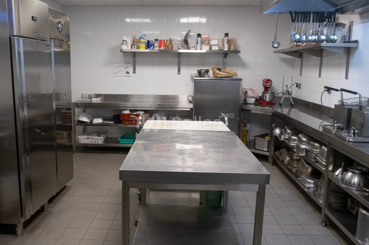 Cuisine professionnelle avec plan de travail en inox, étagères de rangement, réfrigérateur et ustensiles suspendus.