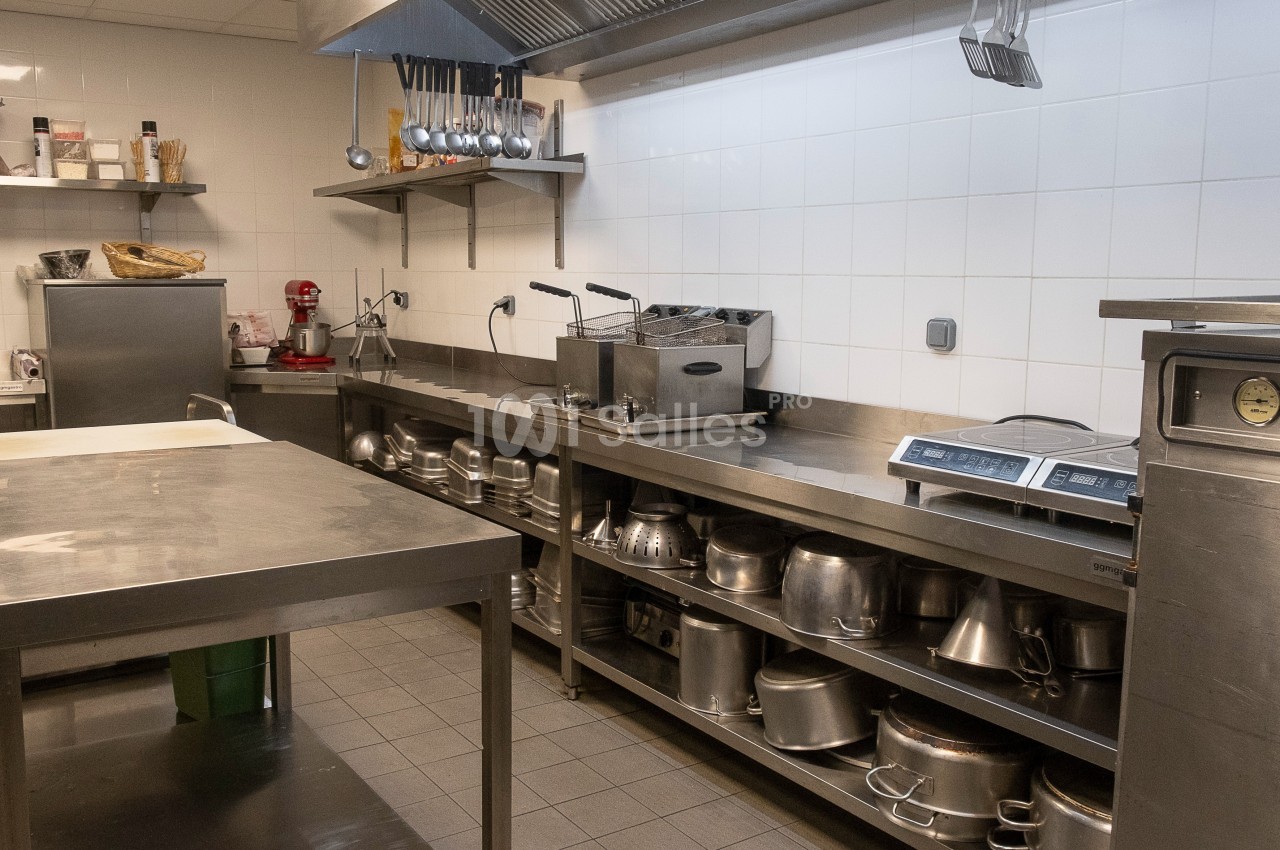 Cuisine professionnelle équipée avec plans de travail en inox, étagères remplies d'ustensiles et appareils de cuisson.