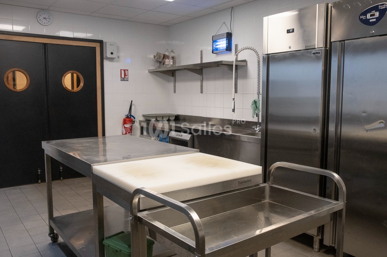 Cuisine professionnelle avec plans de travail en inox, réfrigérateurs, étagères murales et éclairage au plafond.