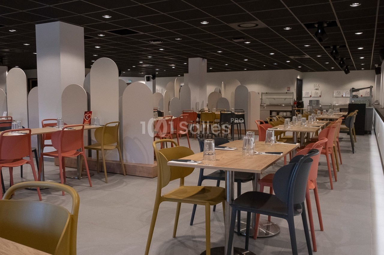 Salle de restaurant moderne avec tables en bois, chaises colorées et séparateurs blancs sous un plafond sombre.