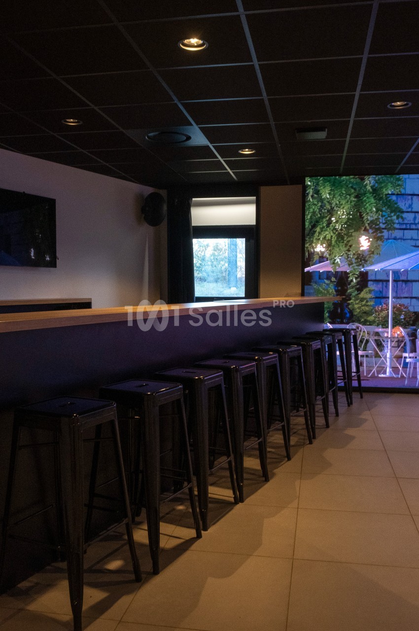 Bar intérieur avec un comptoir en bois, des tabourets hauts noirs alignés et une vue sur une terrasse éclairée.