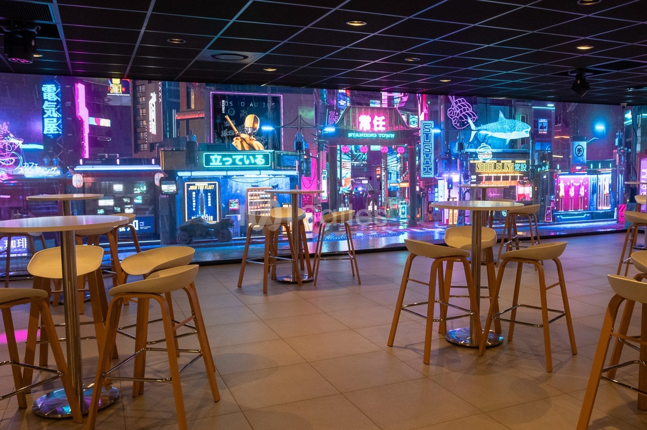 Salle moderne avec tables hautes et tabourets, décorée d'un écran mural affichant une rue animée de style néon futuriste.