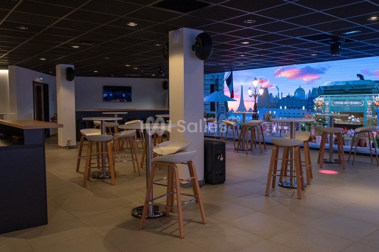 Salle moderne avec tables hautes, tabourets et écran mural affichant une vue urbaine illuminée au crépuscule.