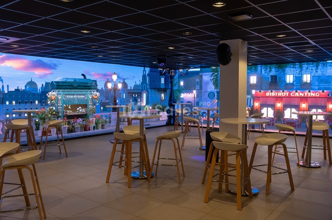 Salle moderne avec tables hautes et tabourets, vue sur une façade illuminée et un décor urbain en arrière-plan.