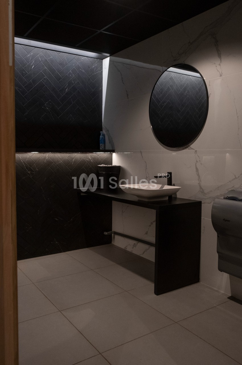 Salle de bain moderne avec lavabo, miroir rond, mur en carrelage noir et blanc, et éclairage tamisé.