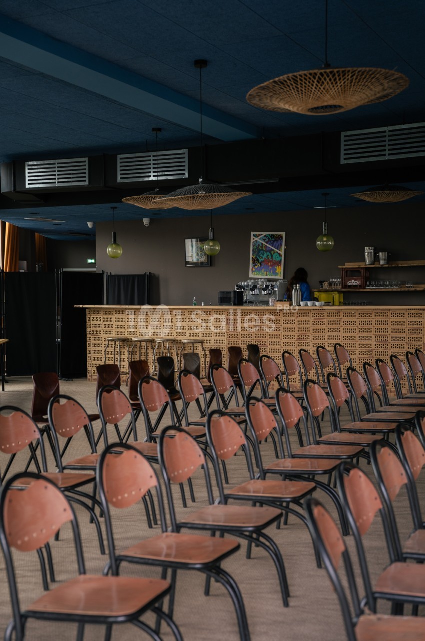 Salle de réception vide avec chaises terracotta en rangées, bar en briques, plafond bleu et luminaires suspendus.
