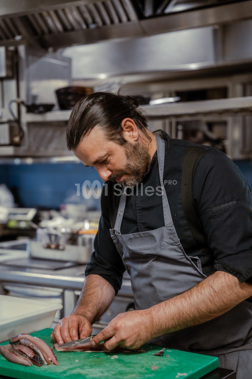 Cuisinier en tablier préparant un poisson sur planche verte en cuisine professionnelle.