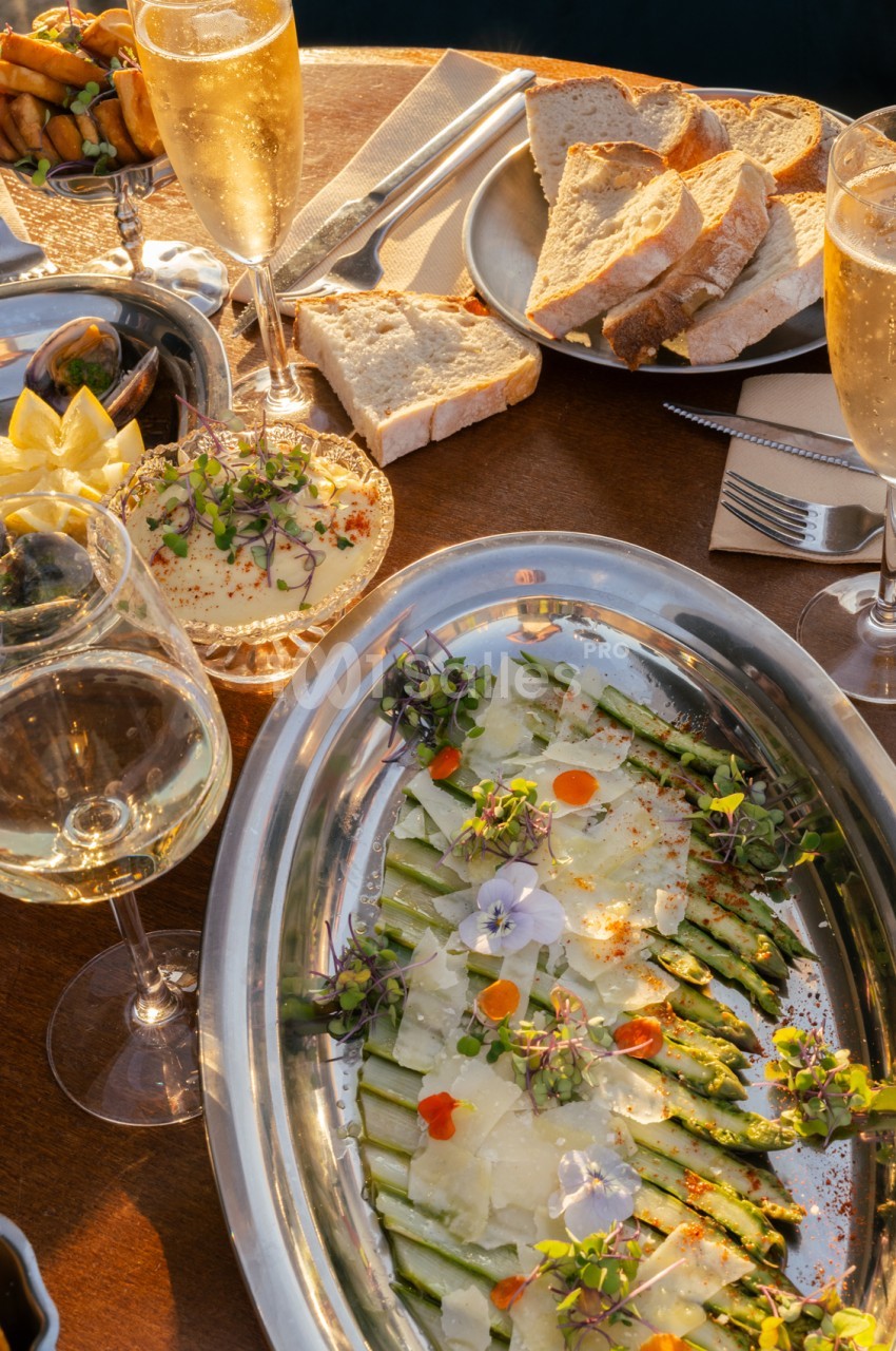 Table dressée en extérieur avec assiette d'asperges sauce blanche et fleurs comestibles, pain grillé, verres de champagne et vin blanc.