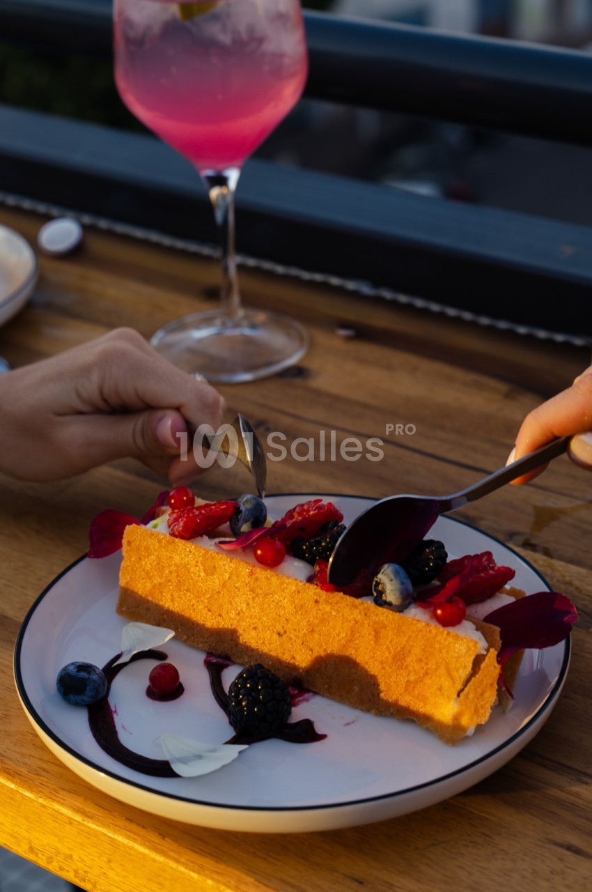 Assiette de dessert avec bâtonnet orange, fruits rouges et baies, accompagnée d'un verre de cocktail rose en terrasse.