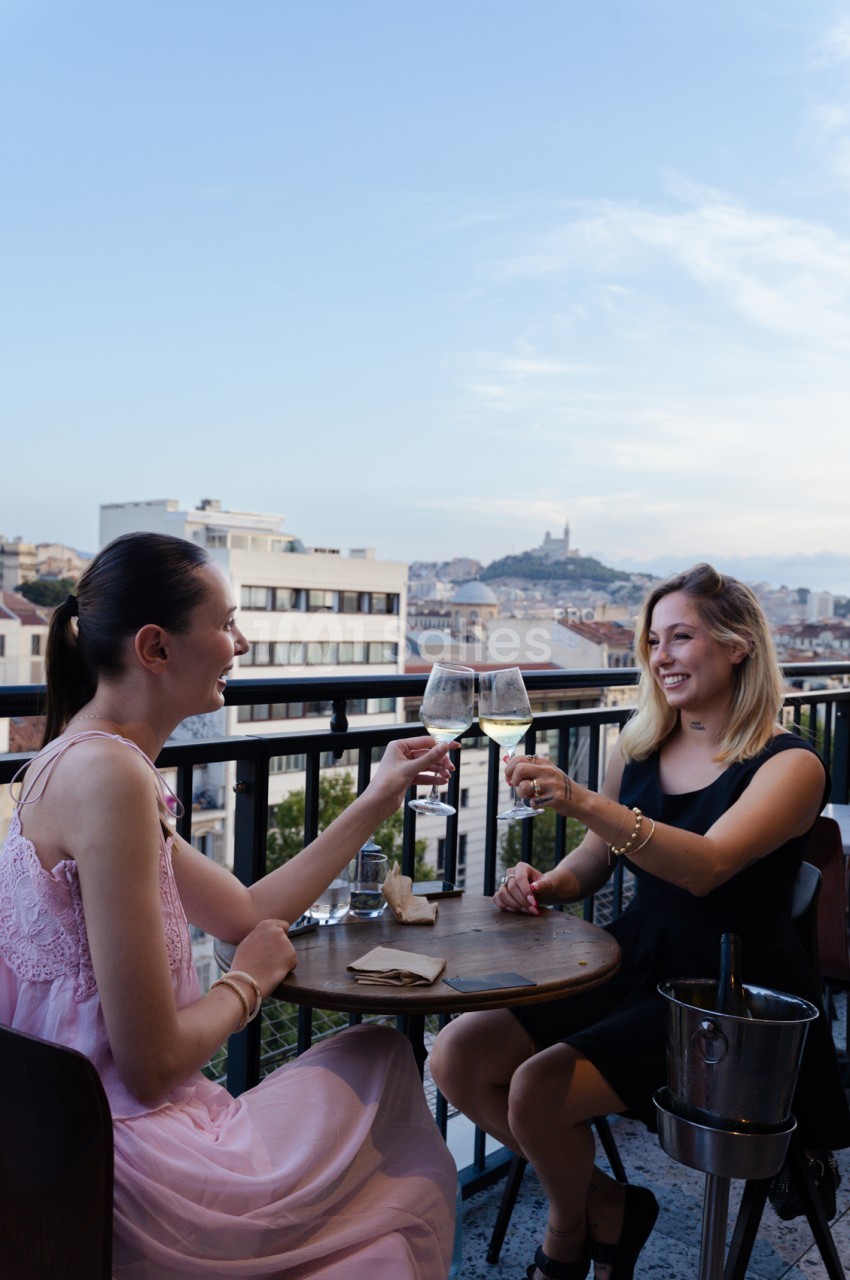 Deux femmes trinquent avec des verres de vin blanc sur une terrasse dominant Marseille et la basilique Notre-Dame-de-la-Garde.