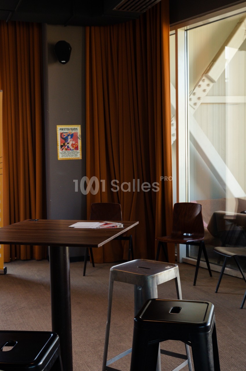 Salle intérieure avec rideaux ocre, table en bois, chaises noires et tabourets métalliques, baie vitrée et affiche murale.