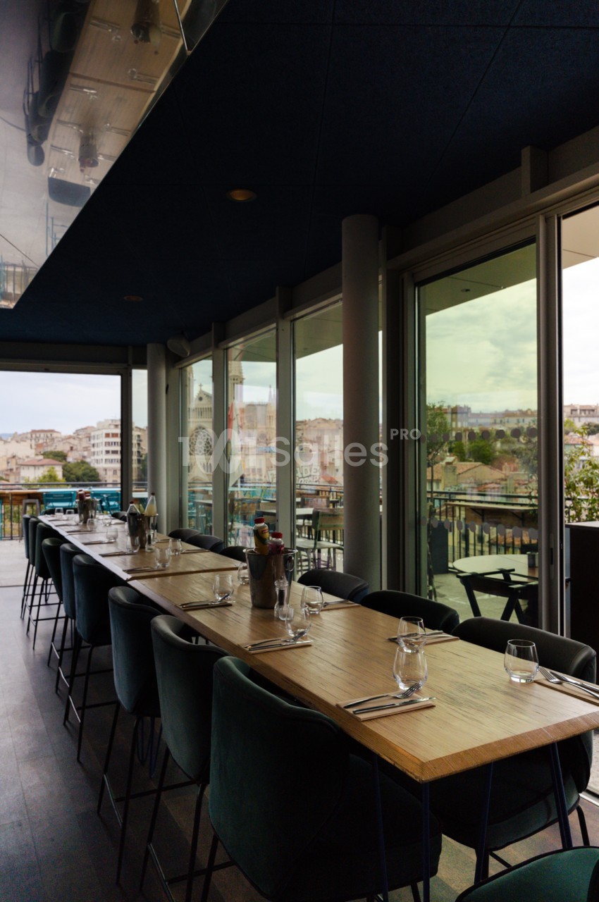 Salle avec tables hautes en bois, chaises noires, baies vitrées ouvertes sur terrasse et vue panoramique urbaine.