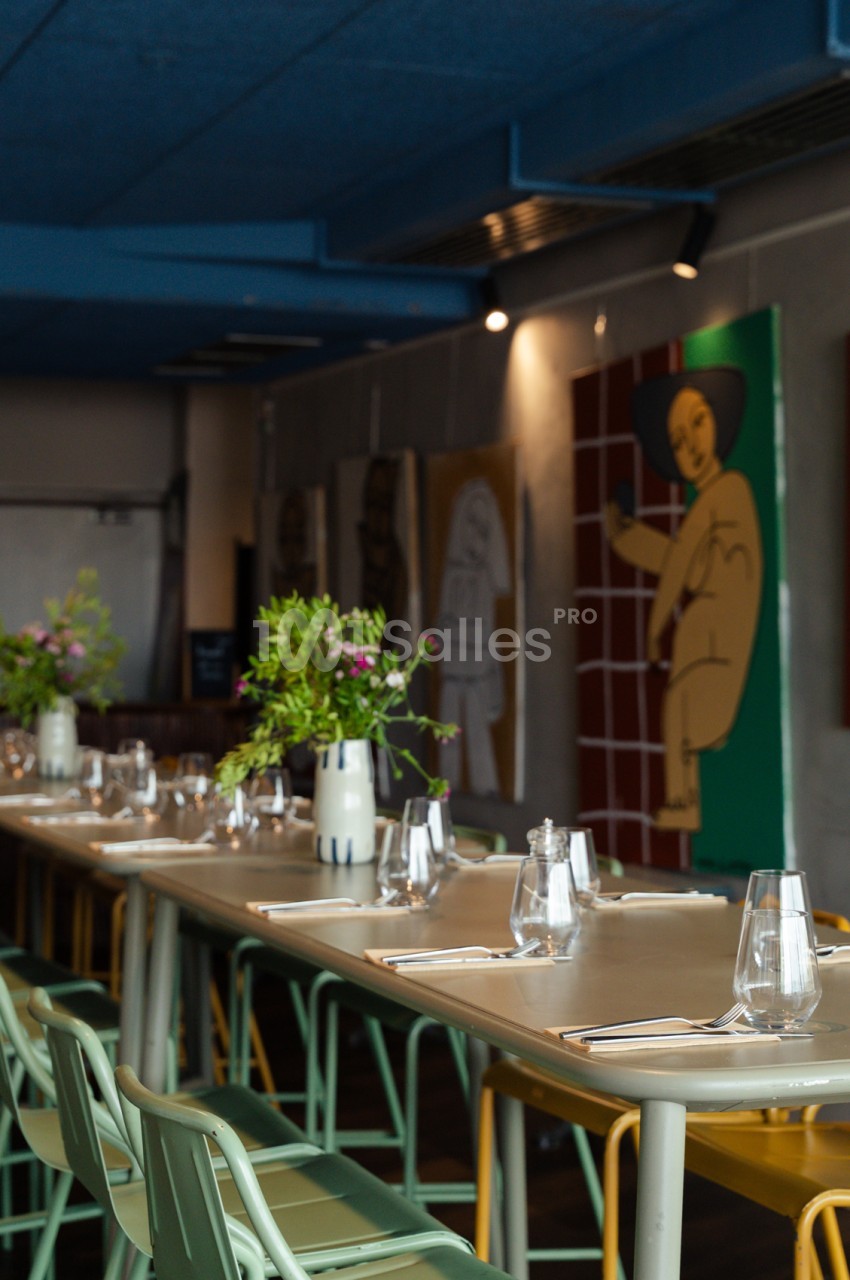 Salle à table longue dressée avec verres et couverts, chaises colorées, plantes en vases, murs gris et artwork vert.