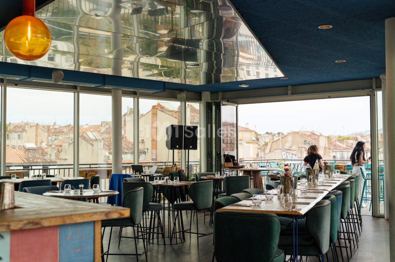 Salle intérieure avec tables hautes, chaises vertes et baies vitrées donnant sur toits marseillais.