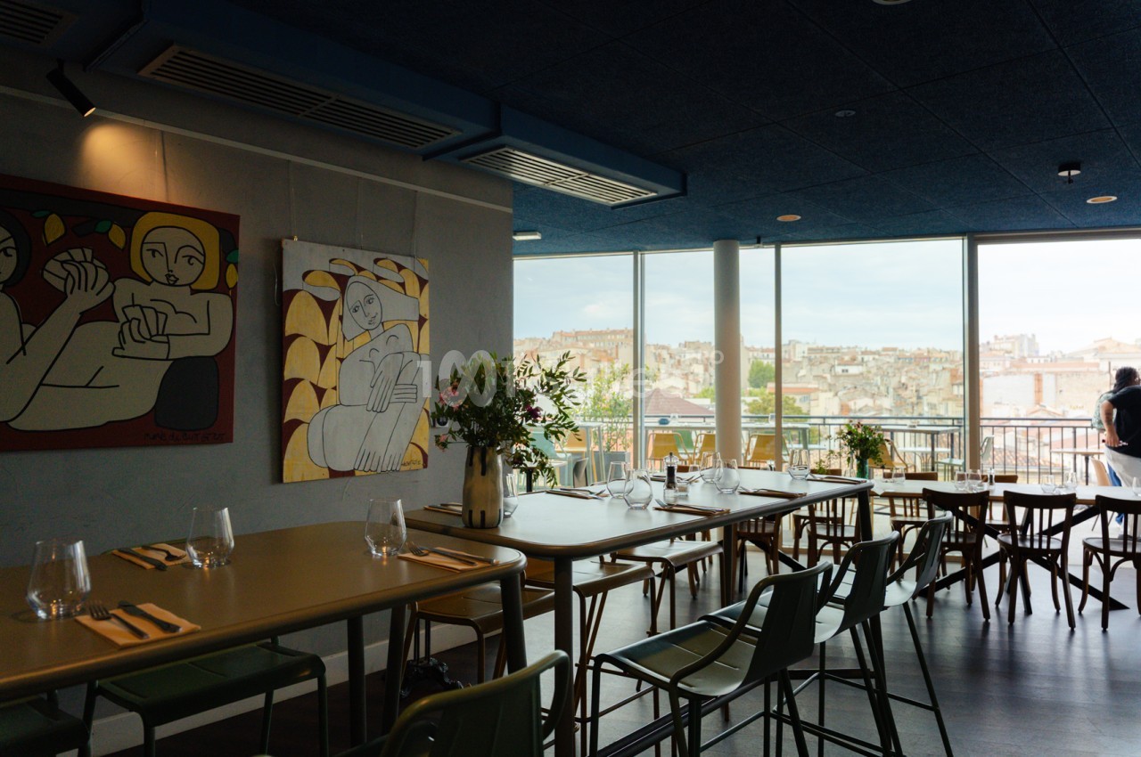Salle de restaurant avec tables dressées, chaises noires, grandes baies vitrées et vue panoramique sur Marseille.