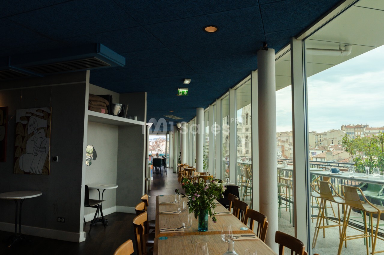 Salle intérieure avec table dressée, chaises en bois, plafond bleu foncé et baies vitrées ouvrant sur terrasse avec vue sur la ville.