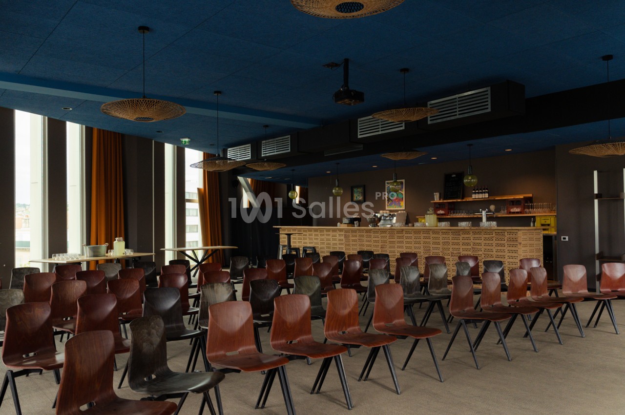 Salle intérieure aux murs bleu et marron avec bar en briques - chaises rouges et noires, lustre doré, baies vitrées.