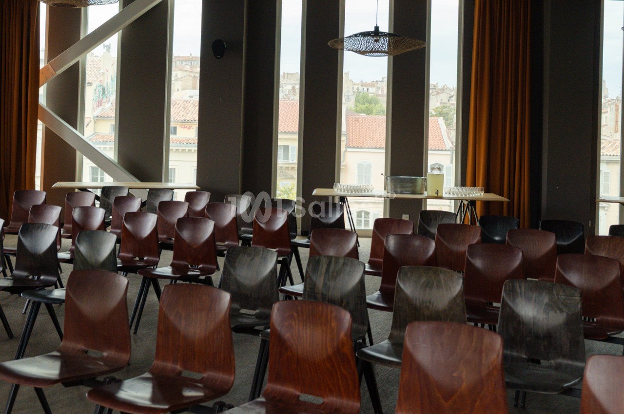 Salle intérieure avec chaises en bois et cuir rouge disposées en rangées, baies vitrées surplombant les toits de Marseille.