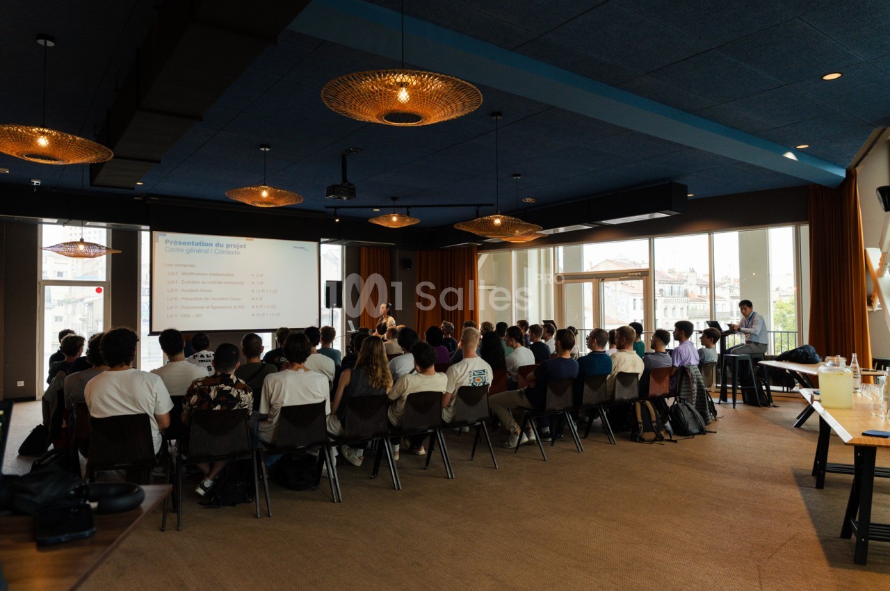 Salle de conférence avec environ 40 participants assis face à un écran de projection - plafond bleu, luminaires dorés, baies vitrées.